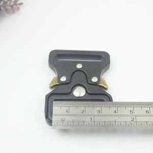 May include: A black metal buckle with gold accents, measuring approximately 1 1/2 inches in length. The buckle is designed for use with webbing or straps.
