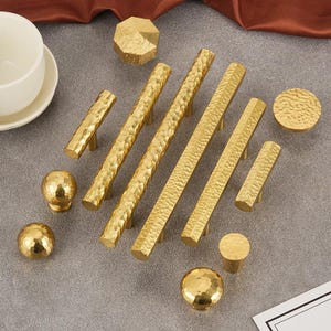 May include: A selection of gold-toned cabinet hardware, including handles and knobs. The hardware has a hammered texture. The set includes long bar handles, short handles, and round knobs, all in a warm, metallic finish. The hardware is suitable for kitchen or furniture projects.