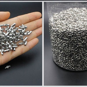 May include: A large quantity of small, silver-colored metal beads in the shape of a capsule. The beads are shown in a hand and in a clear container.