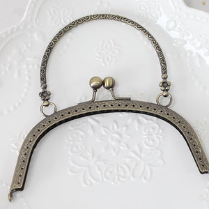 Könnte beinhalten: Ein bronzefarbener Metallrahmen für eine Handtasche mit einem verzierten Design. Der Rahmen hat einen oberen Griff und einen Verschluss.