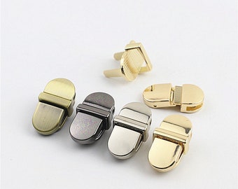 May include: Five metal clasps in different finishes: antique brass, gunmetal, nickel, gold, and brass. The clasps are oval-shaped with a rectangular bar across the center. They are used for securing bags, purses, and other accessories.