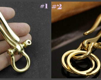 May include: Two gold-toned metal keychains with a hook and a ring. The keychain on the left is held in a hand. The keychain on the right is on a table. The keychain on the left has a screw on the side. The keychain on the right has three rings.