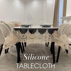 Könnte beinhalten: Eine transparente Silikon-Tischdecke mit beigem Spitzensaum bedeckt einen schwarzen Tisch. Der Tisch ist mit schwarzen Tellern, Weingläsern gedeckt und von beigen Stühlen umgeben. Der Text "Silicone TABLECLOTH" ist abgebildet.