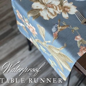 Op de afbeelding: Blauwe waterdichte tafelloper met een bloemen- en kolibrie-ontwerp. De loper heeft grote witte en bruine bloemen, met kleinere roze bloemen en groene bladeren. De tekst "Waterproof TABLE RUNNER" wordt weergegeven.