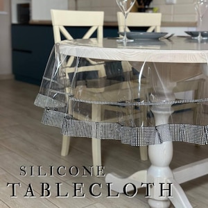 May include: A clear silicone tablecloth with a decorative black and white patterned border. The tablecloth is draped over a round wooden table, with the text "SILICONE TABLECLOTH" at the bottom of the image. Wine glasses and plates are set on the table.