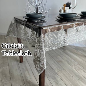 May include: A clear plastic tablecloth with a silver lace design. The tablecloth is on a wooden table with two plates and a glass.