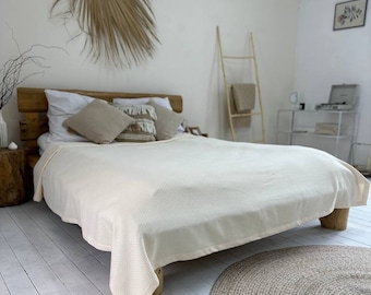 Könnte beinhalten: Ein weißes Bett mit einer weißen Waffeldecke und einem braunen gewebten Teppich auf einem weißen Holzboden. Das Bett hat ein hölzernes Kopfteil mit Kissen und ein getrocknetes Palmblatt hängt an der Wand dahinter.