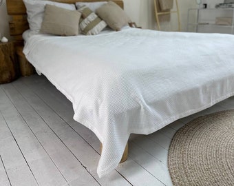 Könnte beinhalten: Ein Schlafzimmer mit einem Bett, das mit einer weißen, strukturierten Decke und beigen Kissen bedeckt ist. Das hölzerne Kopfteil und die neutralen Töne schaffen eine entspannte Atmosphäre. Ein gewebter Teppich und eine Holzleiter ergänzen die Einrichtung.