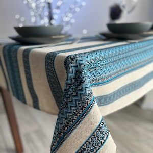 Op de afbeelding: Een wit tafelkleed met blauwe en zwarte geborduurde strepen. Het tafelkleed heeft een geometrisch patroon en is gemaakt van een geweven stof.