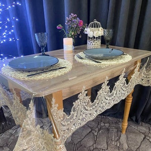 Puede incluir: Una mesa de madera con un mantel de plástico transparente y una superposición de encaje. La mesa está puesta con dos cubiertos, cada uno con un plato azul, cubiertos negros y un vaso. Una pequeña jaula de pájaros blanca con flores blancas se encuentra en el centro de la mesa.