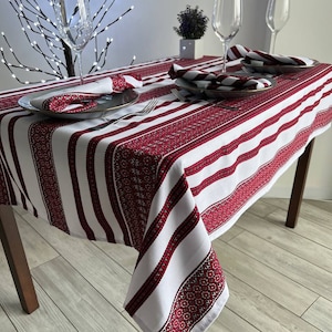 May include: A white tablecloth with red striped and embroidered borders. The table is set with plates, napkins, and wine glasses. The tablecloth has a traditional design, perfect for a special occasion.