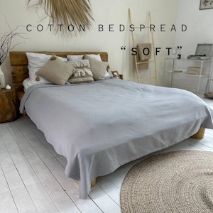 May include: A light gray cotton bedspread with a waffle texture, on a wooden bed frame with a natural wood headboard. The bedspread is on a white wood floor with a brown round rug.