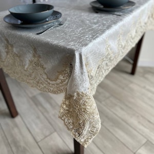 May include: A white tablecloth with a gold lace border. The tablecloth is on a wooden table with two plates and bowls on top.