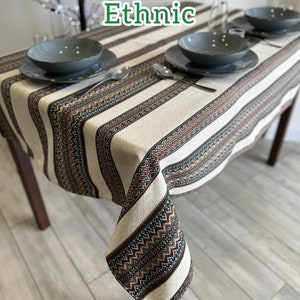 May include: A white tablecloth with brown, green, and red geometric patterns. The tablecloth is laid on a wooden table with two place settings, each with a gray plate and a silver fork.