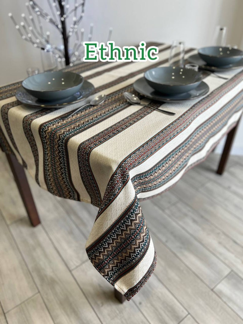 May include: A white tablecloth with brown, green, and red geometric patterns. The tablecloth is laid on a wooden table with two place settings, each with a gray plate and a silver fork.