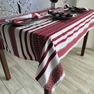 May include: A white and red striped tablecloth with embroidered details. The tablecloth is set for a dinner with two place settings, including plates, napkins, and wine glasses.