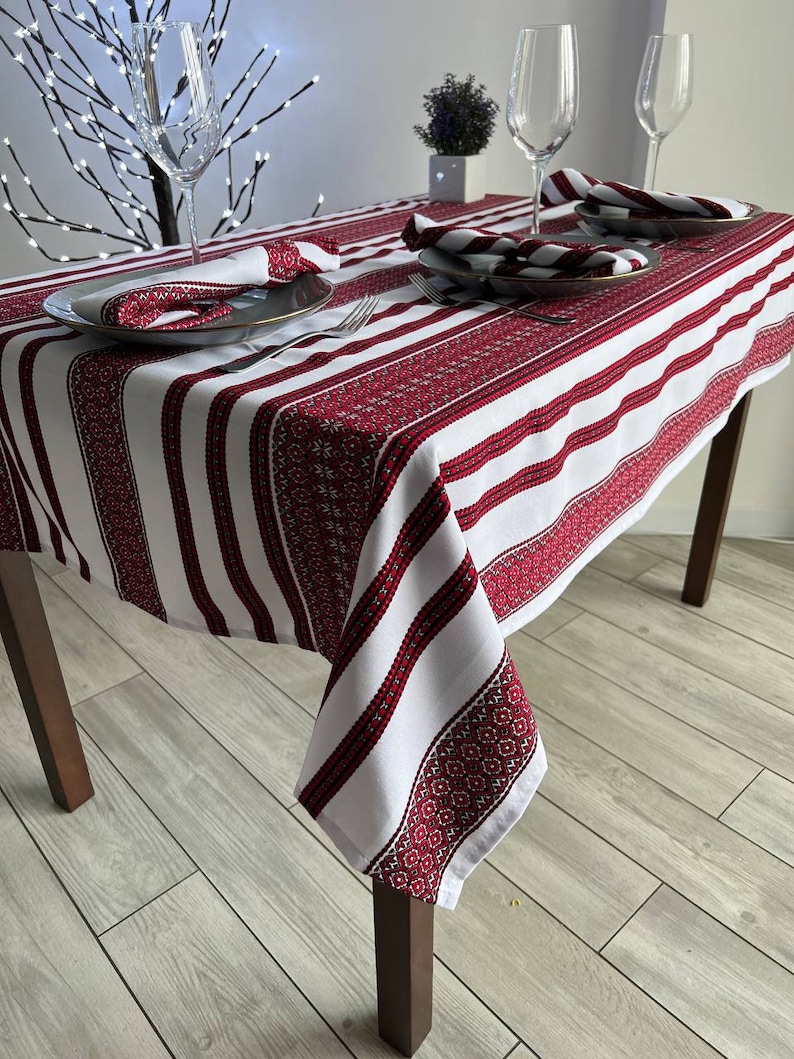 May include: A white and red striped tablecloth with embroidered details. The tablecloth is set for a dinner with two place settings, including plates, napkins, and wine glasses.