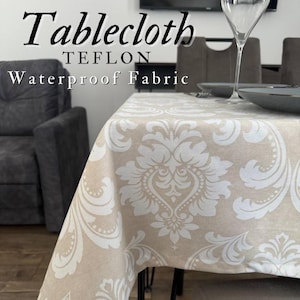 Könnte beinhalten: Beige Tischdecke mit weißem Damastmuster. Die Worte "Tablecloth, Teflon, Waterproof Fabric" sind links oben aufgedruckt. Ein Esstisch ist mit Tellern und einem Weinglas gedeckt.