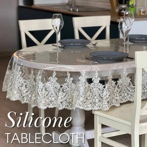 May include: A clear silicone tablecloth with a white lace border covers a round table set for a meal. The tablecloth has the words "Silicone TABLECLOTH" printed on it. The table is set with plates, silverware, and wine glasses.