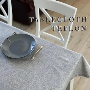 May include: A gray Teflon tablecloth covers a table, with a gray plate and fork set. The text "TABLECLOTH TEFLON" is printed on the image. Two white chairs are in the background.