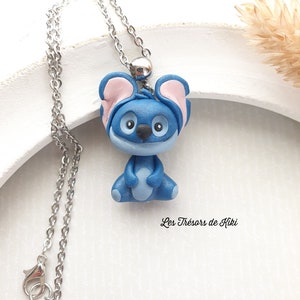 May include: A blue Stitch character pendant necklace with pink ears, a silver chain, and a silver clasp. The necklace is on a white background with the text "Les Trésors de Kiki".