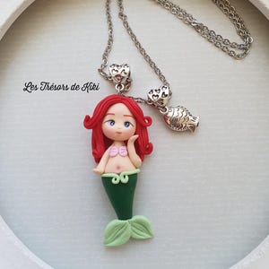 May include: A handmade necklace featuring a mermaid pendant with red hair, a green tail, and a silver chain. The necklace also includes a fish charm and heart-shaped accents. The text "Les Trésors de Kiki" is visible.