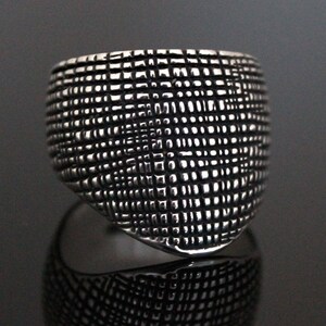 Op de afbeelding: Een zilveren ring met een getextureerd, geometrisch ontwerp. De ring is breed en heeft een afgeronde vorm.