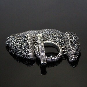 Könnte beinhalten: Ein silbernes Kettenarmband mit einem einzigartigen Toggle-Verschluss. Das Armband besteht aus mehreren Kettensträngen, die jeweils ein anderes Muster aufweisen. Der Verschluss ist eine silberne Stange mit einem Ring an einem Ende und einem Haken am anderen.