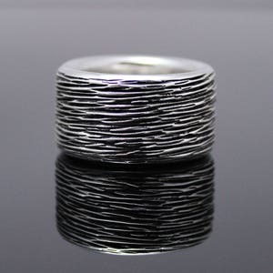 以下が含まれることがあります： テクスチャ加工が施された、畝のあるデザインの銀の指輪。指輪は幅広で、丸みを帯びた形をしています。