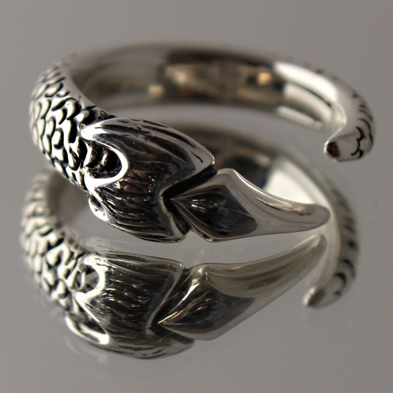 Puede incluir: Un anillo de plata con un dise&ntilde;o detallado de cola de pez. El anillo es abierto y ajustable.