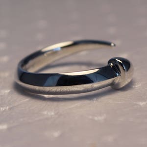 Puede incluir: Un anillo de plata con una banda redonda y lisa y un detalle texturizado elevado en la parte superior.