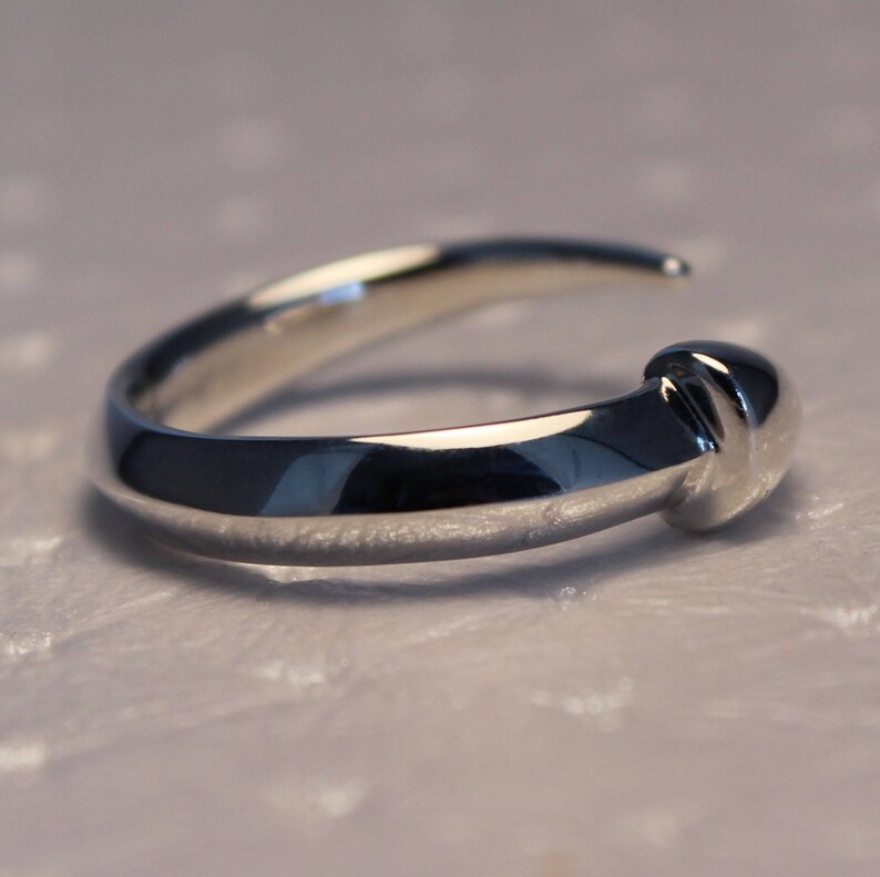 Puede incluir: Un anillo de plata con una banda redonda y lisa y un detalle texturizado elevado en la parte superior.