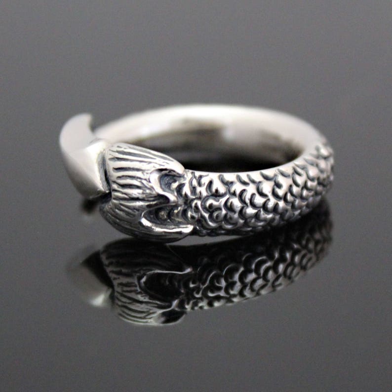 Puede incluir: Un anillo de plata con un dise&ntilde;o detallado de cola de sirena. La cola est&aacute; enroscada alrededor de la banda del anillo, y las escamas son visibles.
