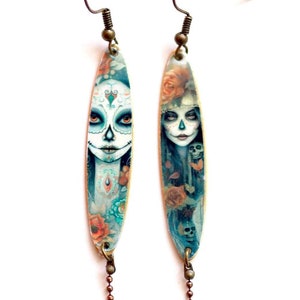 Puede incluir: Un par de pendientes largos y ovalados con ilustraciones del Día de los Muertos. Cada pendiente presenta una cara pintada con detalles florales y de calaveras, con una cadena colgante de pequeñas cuentas. Fondo blanco.