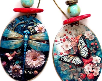 Könnte beinhalten: Ein Paar Hängeohrringe mit ovalen Anhängern, die eine Libelle und einen Schmetterling auf einem floralen Hintergrund zeigen. Die Anhänger sind mit roten und türkisfarbenen Perlen verziert.