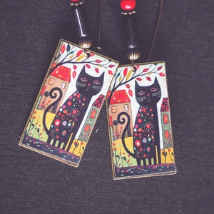 May include: A pair of dangle earrings with black cat illustrations on rectangular metal charms. The cats are black with colorful polka dots and are standing in front of small houses. The earrings have red and black beads on the wire.