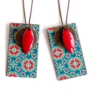 Puede incluir: Un par de pendientes colgantes con un diseño rectangular estampado en tonos azules, verdes y rojos. Los pendientes presentan un dije rojo en forma de lágrima y un dije redondo de color bronce.