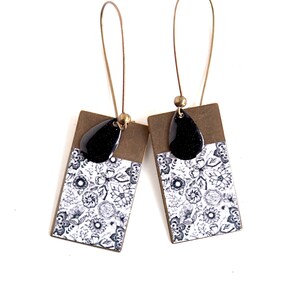 Puede incluir: Un par de pendientes colgantes con un patrón floral blanco y negro en una pieza rectangular de metal. Los pendientes tienen un encanto en forma de lágrima negra que cuelga de la parte superior del rectángulo.