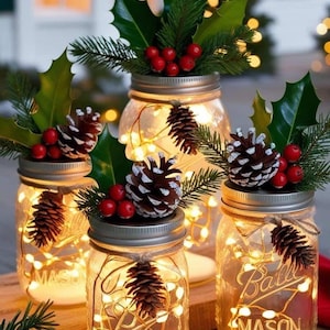 Może przedstawiać: Słoiki Masona ozdobione lampkami choinkowymi, szyszkami, ostrokrzewem i czerwonymi jagodami. Słoiki ustawione są na drewnianej powierzchni, tworząc ciepłą, świąteczną atmosferę. Na słoikach widnieje napis "MASON".