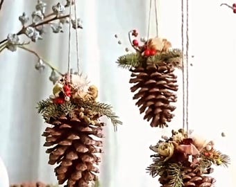 Puede incluir: Tres adornos colgantes de piñas. Cada piña marrón está adornada con vegetación, bayas y flores secas, suspendidas por hilo. Un lazo rojo y ramas con bayas rojas se suman al tema festivo navideño.