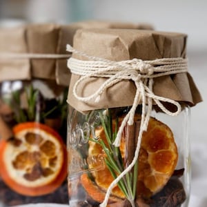 Puede incluir: Dos frascos de vidrio transparente llenos de rodajas de naranja secas, palitos de canela, anís estrellado y ramitas de romero. Los frascos están sellados con papel marrón y atados con hilo.