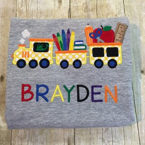 Può includere: Una maglietta grigia con un treno di autobus scolastico giallo ricamato su di essa. Il treno trasporta materiale scolastico come matite colorate, matite, libri e una mela. Il nome "BRAYDEN" è ricamato sotto il treno in colori arcobaleno.