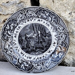 Peut inclure: Une assiette décorative en noir et blanc avec une illustration détaillée d'une scène de duel au centre. L'assiette présente des motifs floraux et d'oiseaux ornés sur le bord, avec le texte "DUEL A L'EPEE" en bas.