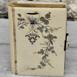 Peut inclure: Un petit livre rectangulaire blanc cassé avec une couverture à charnière. Le devant présente une illustration à l'encre noire d'un calice entouré de vignes florales et de petits accents en nacre incrustés. Un loquet en métal sombre est fixé sur le côté.