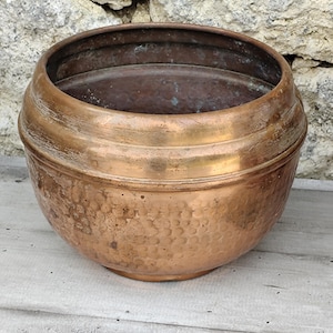 Peut inclure: Un pot en cuivre rond et antique avec une texture martelée sur la moitié inférieure. Le pot a une forme large et arrondie avec une bande décorative au milieu. Le cuivre a une patine chaude et vieillie, montrant des signes d'utilisation.