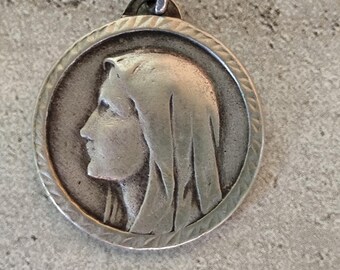 May include: A silver-toned pendant featuring a profile of a woman with a draped head covering. The round pendant has a detailed border and a small loop for a chain. The image is set against a textured, gray background.