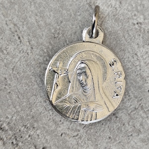 May include: A silver-colored pendant with a detailed image of a religious figure, possibly a saint, holding a cross. The pendant has the inscription "S. RITA" around the figure's head and a small loop for a chain.