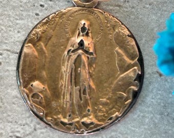 Puede incluir: Un colgante redondo de color bronce con un lazo dorado. El colgante presenta un relieve detallado de una figura religiosa con texto alrededor del borde. El colgante tiene un borde oscuro y está sobre un fondo gris.