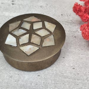 Peut inclure: Une boîte ronde en laiton antique avec une incrustation géométrique en nacre sur le couvercle. La boîte est posée sur un fond gris avec une branche de fleurs rouges. Un objet de rangement décoratif.