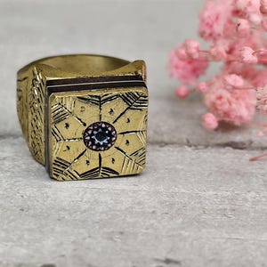 Può includere: Un anello dorato con una faccia quadrata caratterizzata da un intricato design geometrico e una pietra centrale multicolore. Anche il gambo dell'anello è strutturato. Lo sfondo include fiori rosa.